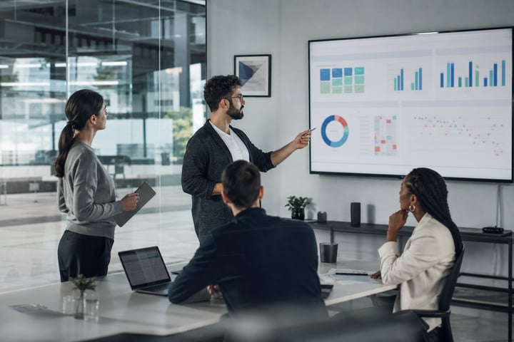 Consultant presenting a CMDB health dashboard with completeness, correctness and compliance metrics to a team in a meeting room