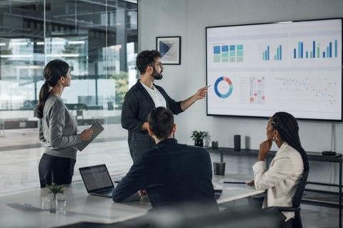 Consultant presenting a CMDB health dashboard with completeness, correctness and compliance metrics to a team in a meeting room
