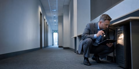 Man in a grey suit crouching in an office corridor, shining a torch on an old server tucked under a desk