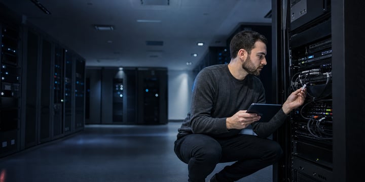 Network engineer crouching beside an equipment rack in a dark server room, checking a cable while holding a tablet