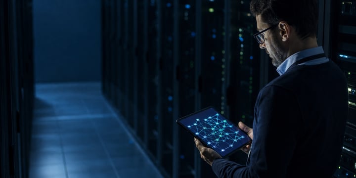 A security analyst holds a tablet showing a glowing network map in a dim data centre server aisle.